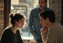 Photo of An Emotional Conversation Unfolds in a Quiet Café as an Unexpected Visitor Pauses to Listen at the Table